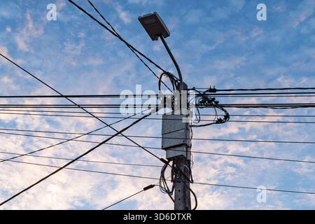 Une photo à angle bas d'un poteau utilitaire en béton avec un désordre chaotique de fils enchevêtrés, de boîtes et d'un lampadaire contre un ciel bleu lumineux et nuageux. Banque D'Images