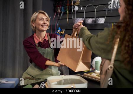 Femme d'âge moyen caucasien souriant tout en remettant un sac à provisions en papier à la jeune femme adulte dans un magasin de fleurs, comptoir affichant des fournitures florales et des matériaux d'emballage en arrière-plan Banque D'Images