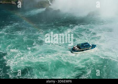 Vue de dessus du bateau d'excursion brave les eaux tourbillonnantes et mousseuses à la base des chutes du Niagara, au milieu de la silhouette brumeuse des chutes en arrière-plan, symb Banque D'Images