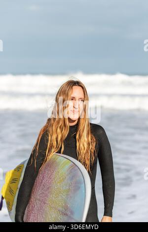 Portrait d'une femme surfeuse en combinaison debout sur une mer mouillée avec planche de surf près de la mer dans la zone côtière le jour d'été Banque D'Images