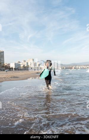 Jeune femme de surf attentionnés en combinaison avec planche de surf qui s'étourne sur la mer lavée par la mer Banque D'Images