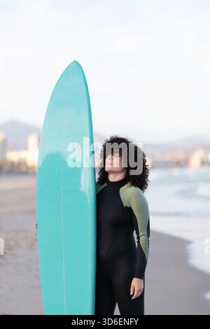 Jeune femme de surf attentionnée en combinaison avec planche de surf debout avec les yeux fermés sur le bord de mer lavé par la mer Banque D'Images