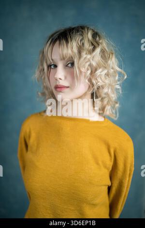 Une jeune femme aux cheveux blonds bouclés pose sur un fond bleu, portant un haut jaune moutarde. Son expression confiante et sa tenue décontractée créent un Banque D'Images