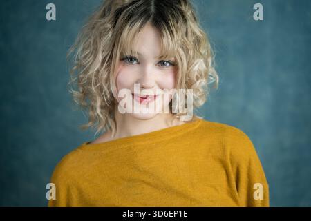 Une jeune femme souriante aux cheveux blonds bouclés et à la frange porte un pull jaune moutarde. Elle se tient debout sur un fond bleu, exsudant chaleur et con Banque D'Images
