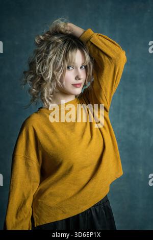 Jeune femme aux cheveux blonds bouclés pose sur un fond bleu texturé, portant un pull jaune. Le contraste des couleurs ajoute de la profondeur et de la chaleur à Banque D'Images