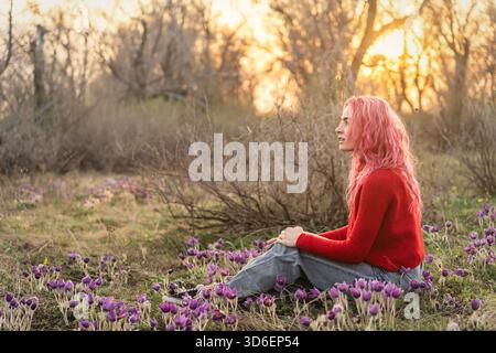 Une jeune femme aux cheveux roses éclatants est assise paisiblement parmi les fleurs en fleurs dans un cadre naturel. La lumière chaude du soleil crée un atmo serein et coloré Banque D'Images