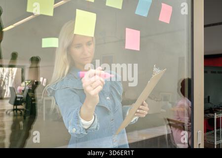 Travailleur féminin concentré debout avec une planchette à pince sur le lieu de travail et écrivant sur un mur de verre avec des notes adhésives colorées Banque D'Images