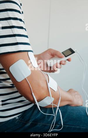 l'invention concerne un homme tenant un dispositif d'électrothérapie portable tout en utilisant des tampons adhésifs sur son bras pour un traitement de stimulation musculaire à l'intérieur Banque D'Images