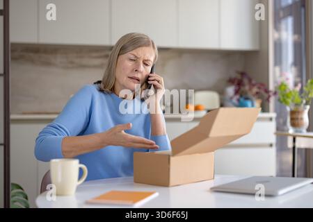 Femme mature bouleversée à la table de cuisine à la maison se plaignant sur smartphone d'un colis incorrect reçu après déballage, exprimant frustration et insatisfaction Banque D'Images