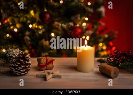 Bougie allumée entourée de pommes de pin, de petits cadeaux et d'un décor festif créant une atmosphère chaleureuse de Noël avec des lumières d'arbre floues en arrière-plan Banque D'Images
