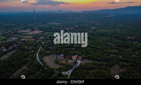 Village rural et vaste forêt dispersée sous un coucher de soleil, contrasté par une rangée lointaine d'éoliennes modernes à l'horizon Banque D'Images