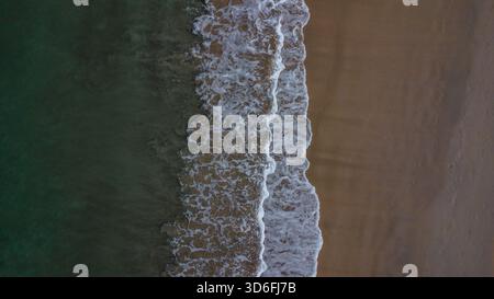 Des vagues blanches mousseuses se brisent entre le sable sombre et l'eau profonde de l'océan vert émeraude Banque D'Images
