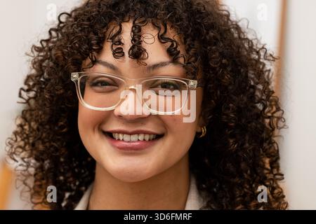 Femme souriant tout en portant des lunettes à cadre clair et de petites boucles d'oreilles en or dans le portrait de bureau Banque D'Images