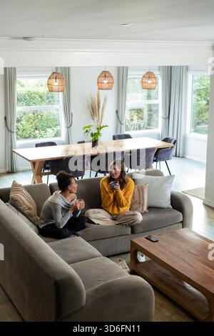 Diverses amies féminines sirotant du café dans des tasses sur un canapé gris dans le salon avec des oreillers jetés. Confort, compagnie, modernité, confort, relaxatio Banque D'Images