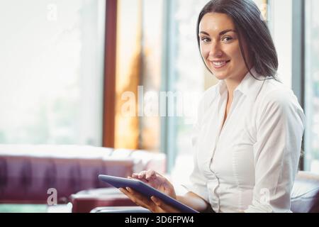 Femme adulte assise sur un canapé dans un salon lumineux portant une blouse blanche tapissant la tablette, espace de copie. Personne, écran tactile, appareil, professionnel, moderne, abic Banque D'Images