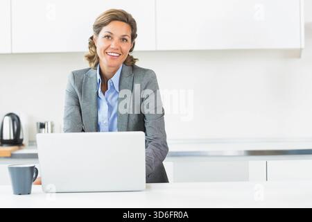 Femme d'âge moyen dans la tenue d'affaires debout au comptoir de cuisine en utilisant un ordinateur portable argenté avec tasse sombre. Professionnel, moderne, minimal, lumineux, espace de travail, Banque D'Images