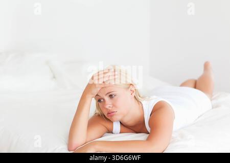Femme couchée sur le lit portant des vêtements de nuit blancs, soutenant la tête avec la main, regardant vers le bas dans la chambre. Minimaliste, serein, contemplatif, solitaire, matelas, l Banque D'Images