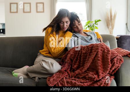 Partenaires féminins divers assis sur le canapé gris embrassant sous une couverture de couleur rouille dans le salon Banque D'Images