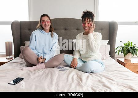 Amies féminines diverses appliquant des patchs de gel sous les yeux tout en étant assis les jambes croisées sur un lit king-size Banque D'Images