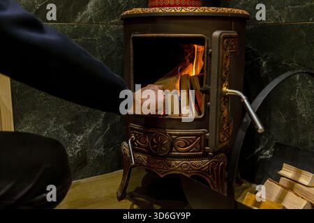 Homme chargeant du bois de chauffage dans le poêle à bois vintage. Gros plan d'une personne plaçant du bois de chauffage dans un poêle à bois en fonte brûlant avec des motifs complexes, pour Banque D'Images
