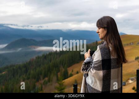 Femme enveloppée dans une couverture profitant de Mountain View. Vue de côté d'une femme tenant une tasse et admirant un paysage de montagne brumeux, enveloppé dans un plaid Banque D'Images