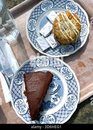 Tranche de gâteau au chocolat riche et grand scone avec filet blanc servi sur des assiettes traditionnelles à motifs avec des portions de beurre sur un plateau en bois uk Banque D'Images