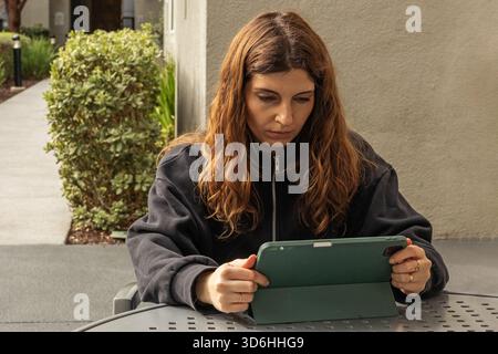 Une femme aux cheveux longs est assise à une table extérieure, concentrée sur sa tablette. Banque D'Images