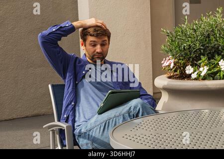 Un homme est assis à une table extérieure, paraissant perplexe tout en tenant une tablette. Banque D'Images