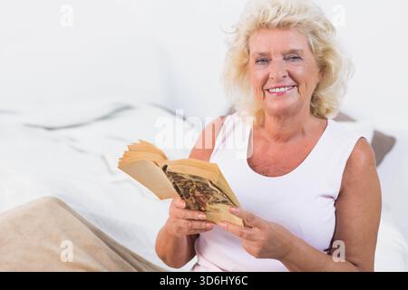 Femme senior assise sur le lit avec une couverture beige portant le dessus tenant le livre de poche, souriant, espace de copie. Lecture, livre, mature, confortable, calme, lumière douce, maison Banque D'Images