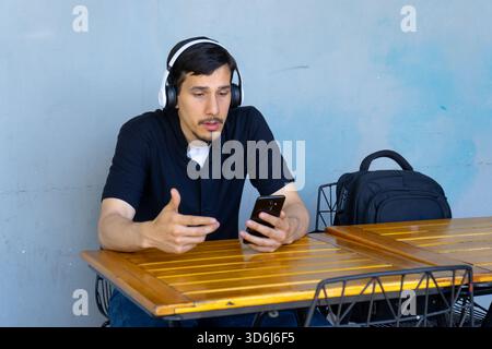 Homme portant des écouteurs et parlant sur smartphone à une table, s'engageant dans le travail à distance ou la conversation en ligne Banque D'Images