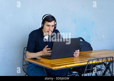 Jeune homme portant des écouteurs et utilisant un ordinateur portable pour l'apprentissage en ligne ou le travail à distance, assis à une table de café Banque D'Images