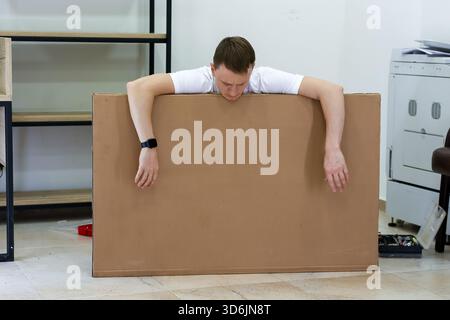 Homme ennuyé flouing avec les bras sur le panneau de carton à l'intérieur. Jeune homme ayant l'air ennuyé et fatigué, affaissant avec les bras suspendus au-dessus de la grande feuille de carton dedans Banque D'Images