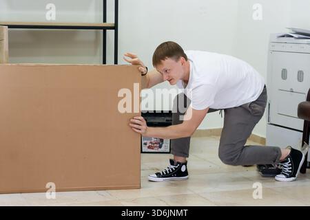 Homme luttant pour soulever une grande boîte en carton à l'intérieur. Jeune homme dans des vêtements décontractés s'efforçant de déplacer une lourde boîte en carton lors de l'assemblage de meubles dans un Banque D'Images