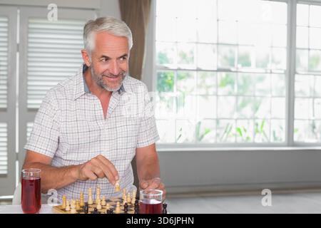 Homme senior portant chemise à carreaux jouant aux échecs sur échiquier en bois à table avec deux grandes lunettes. Ensoleillé, verdure, fenêtre, volets, intérieur, rela Banque D'Images