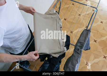 Vue de haut en bas d'un homme en chemise blanche arrangeant un pantalon beige sur un séchoir à vêtements en métal au-dessus du parquet en bois avec d'autres vêtements suspendus à proximité Banque D'Images