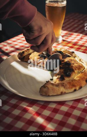Tranchant à la main un calzone cuit sur une assiette blanche à l'aide d'un coupe-pizza, avec un verre de bière sur une nappe à carreaux, gros plan. Banque D'Images