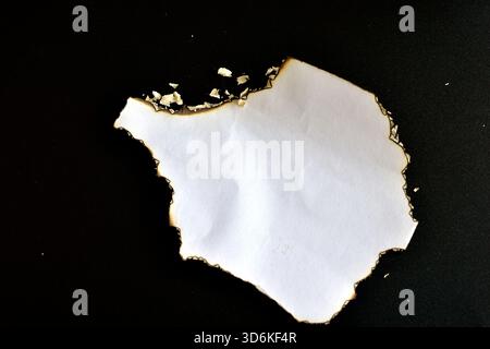 Une feuille de papier avec des bords brûlés et de la cendre repose sur un fond noir. Banque D'Images