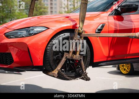 Gros plan d'une voiture rouge soulevée par une dépanneuse avec une grue. Aile avant, roue et pneu de BMW sportive dans l'air sécurisé par un équipement robuste et usé Banque D'Images