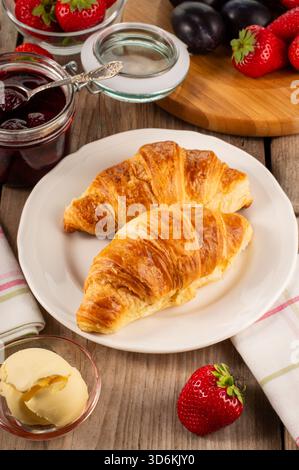 Deux croissants français sur une assiette blanche au premier plan avec de la confiture de fraises et un petit bol de beurre derrière eux sur une table en bois, créant un chaud Banque D'Images