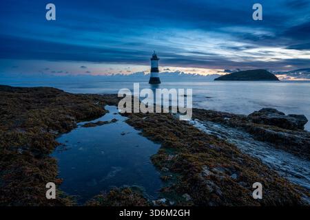 Lumière du matin au-dessus du phare de Penmon point avec des rochers couverts d'algues au premier plan et Puffin Island au-delà sous un ciel en couches Banque D'Images