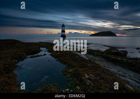 Lumière du matin au-dessus du phare de Penmon point avec des rochers couverts d'algues au premier plan et Puffin Island au-delà sous un ciel en couches Banque D'Images