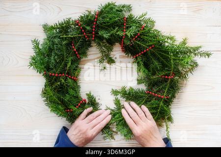 Mains faisant une couronne de l'Avent à partir de branches à feuilles persistantes et une chaîne rouge sur une table en bois clair, décoration pour la saison de Noël, vue de dessus d'Abov Banque D'Images