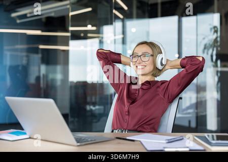 Jeune femme dans un fauteuil de bureau souriant avec des écouteurs sans fil, se penchant en arrière à côté de son ordinateur portable pendant une pause de travail détendue, profitant de la musique et d'un moment de bien-être calme Banque D'Images