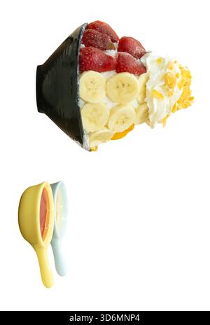 Un ensemble de Bingsu coréen dans un petit bol noir avec plusieurs nappages de fruits et une sauce rouge avec du lait condensé sucré dans des mini cuillères sont isolés sur blanc Banque D'Images