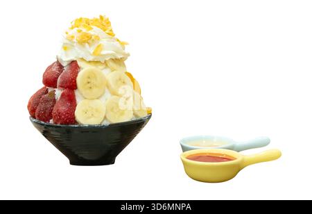 Un ensemble de Bingsu coréen dans un petit bol noir avec plusieurs nappages de fruits et une sauce rouge avec du lait condensé sucré dans des mini cuillères sont isolés sur blanc Banque D'Images