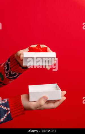 Mains tenant une boîte cadeau blanche ouverte avec un arc rouge sur un fond rouge vif, parfait pour les promotions des fêtes, les célébrations et le marketing festif vi Banque D'Images