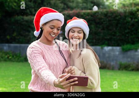 Diverses amies féminines embrassant sur la pelouse dans des chandails et des chapeaux de Père Noël capturant selfie avec smartphone. Festif, amitié, célébration, loisirs, plein air Banque D'Images