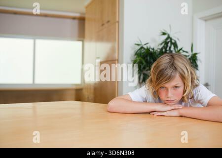 Enfant mâle en t-shirt blanc appuyé sur une table en bois dans la cuisine, avec plante en pot, espace copie. Garçon, contemplatif, solitude, introspection, réfléchi Banque D'Images