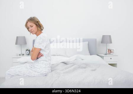 Femme adulte mature assise sur le bord du lit, portant un pyjama à motifs, à côté du réveil, espace copie. Contemplatif, minimaliste, chambre à coucher, neutre, serein Banque D'Images