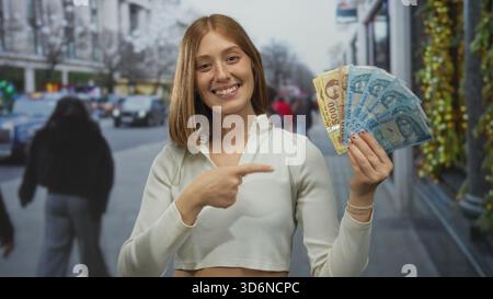 Jeune femme blonde tenant des billets de banque hongrois en forint souriant dans une rue urbaine avec un paysage urbain en arrière-plan, mettant en valeur la monnaie à l'extérieur. Banque D'Images